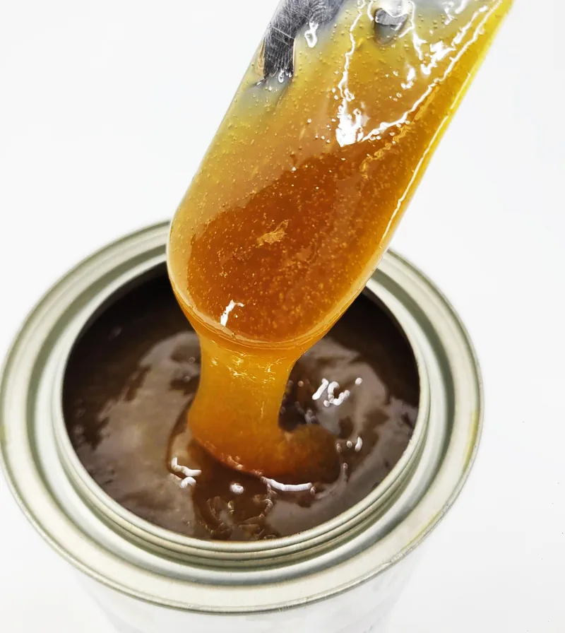 A blade lifts amber open-gear lubricant from a tin, drawing a long string.