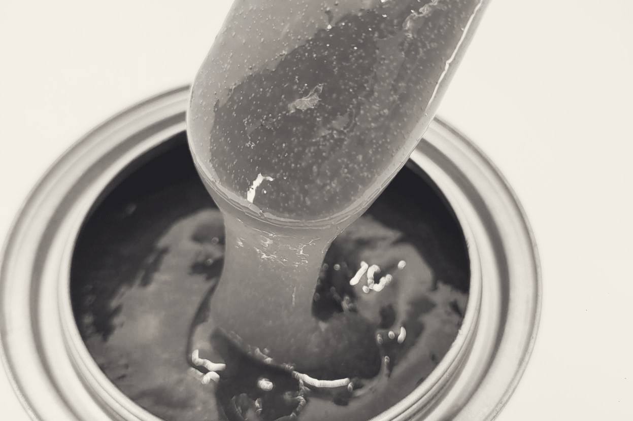 A spatula lifts a thick rope of high-viscosity lubricant out of an open can. The lubricant draws into a long unbroken string before falling back to the darker product below. Fine bubbles are suspended in it.